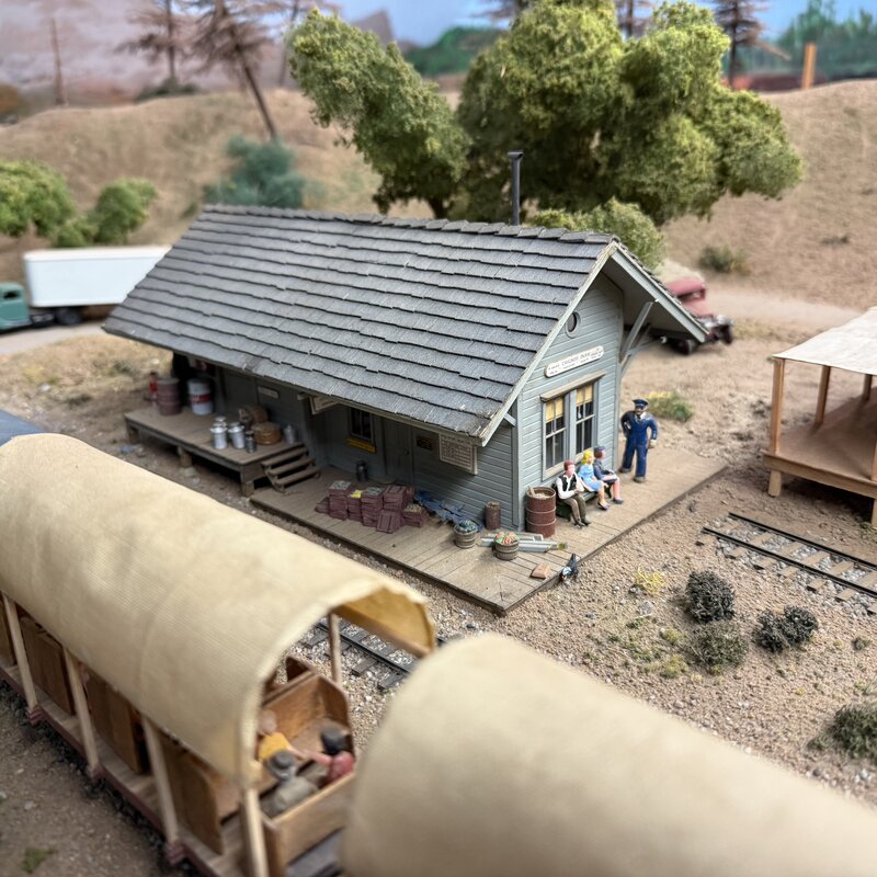 Small trackside building with barrels, crates, seated figures, and covered passenger cars
