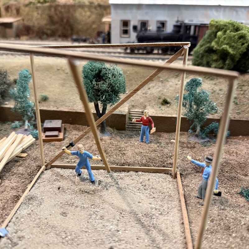 Miniature carpenters assembling a timber frame beside the railroad