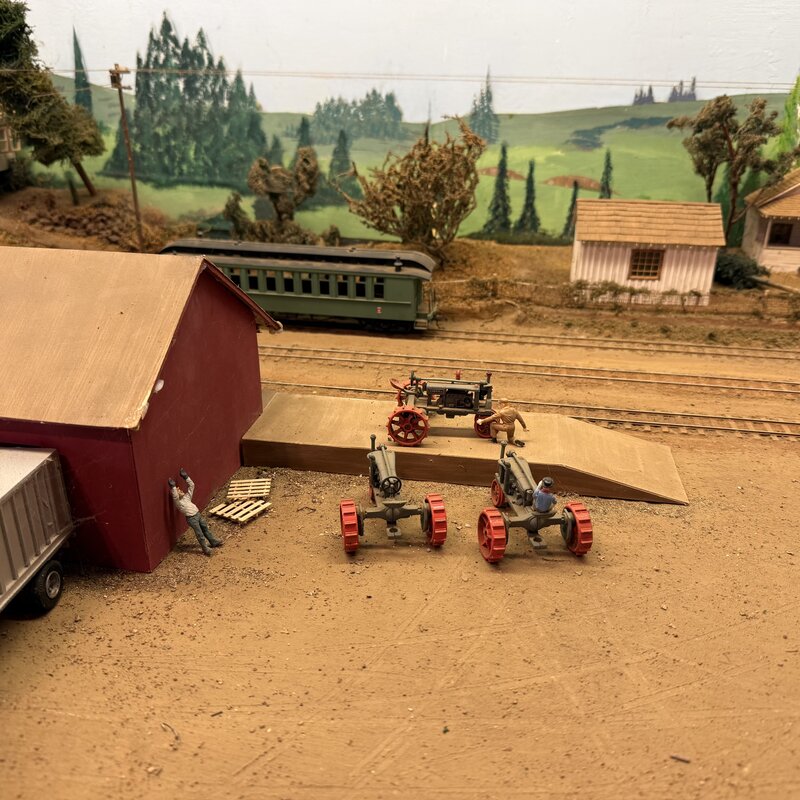 Vintage tractors beside a loading ramp, red shed, and passenger coach