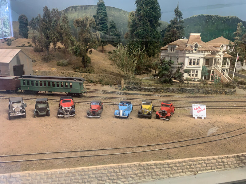 Vintage model cars lined up beside a green passenger coach and Victorian-style house